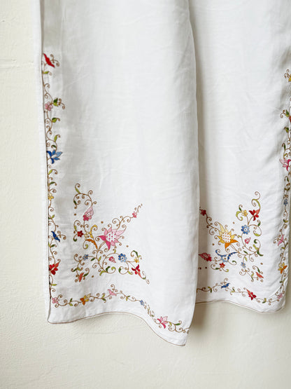 Hem detail of white overalls with colourful floral embroidery hanging on a wall.