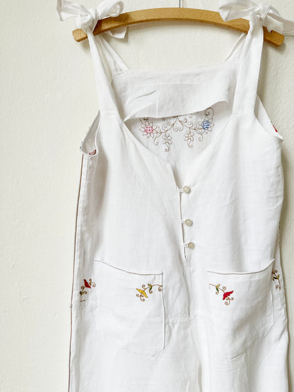 Back detail of White jumpsuit with colourful floral embroidery and button closure hanging on a wall.