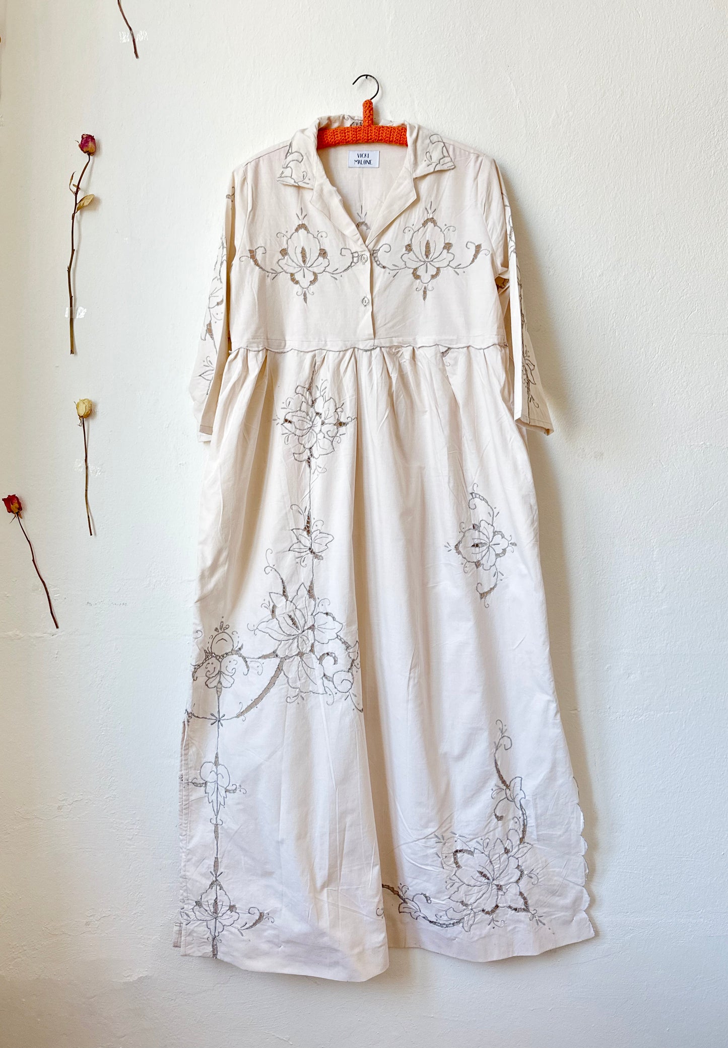 The front view of a maxi length grey embroidered dress hanging on a wall.