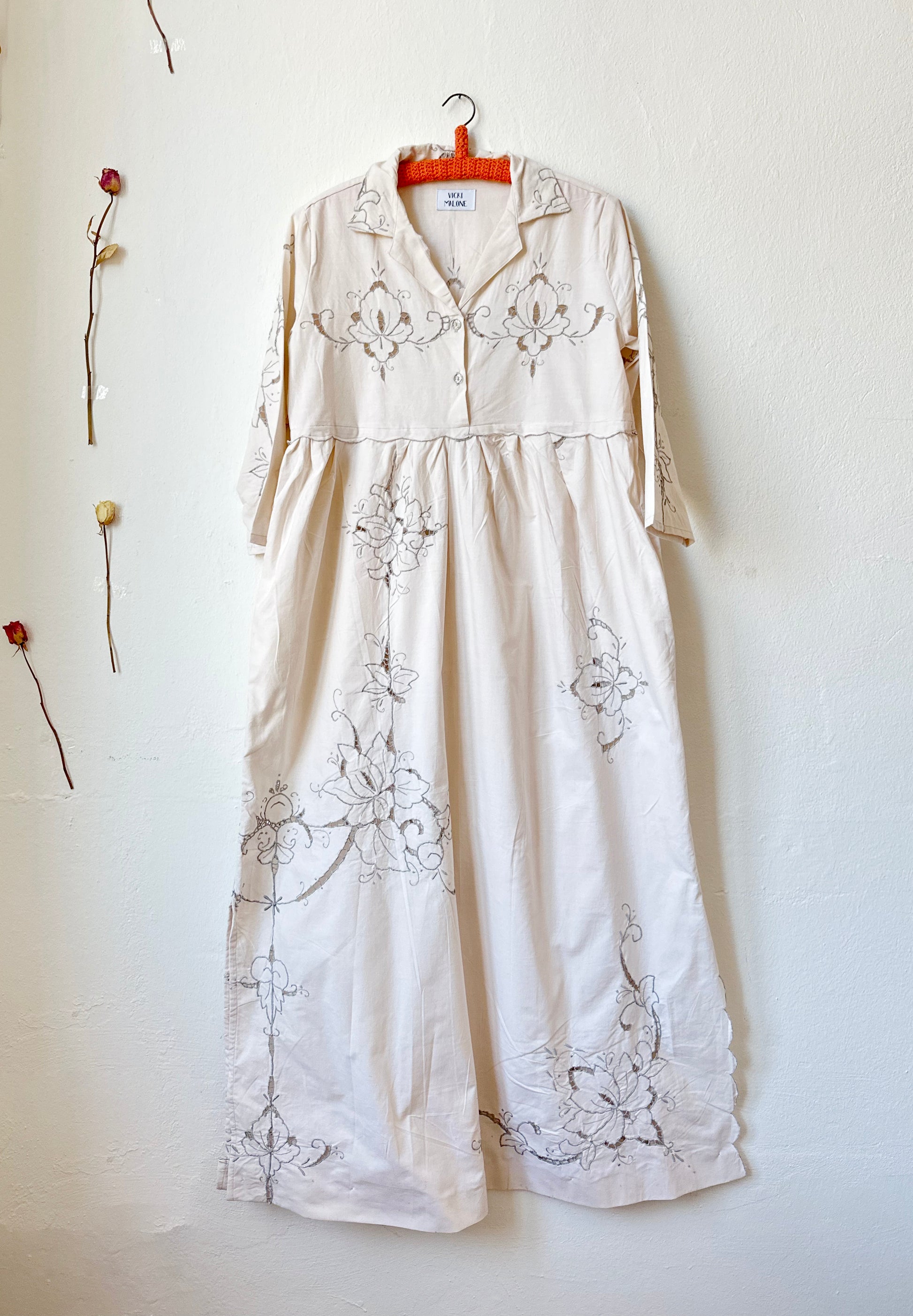 The front view of a maxi length grey embroidered dress hanging on a wall.