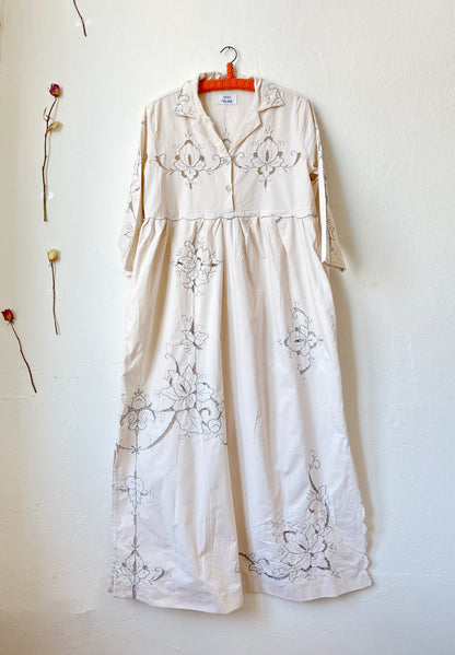 The front view of a maxi length grey embroidered dress hanging on a wall.