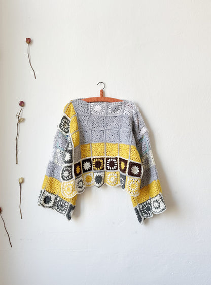 The front view of a crocheted sweater hanging on a white wall. The sweater is grey and yellow made from a granny square blanket.