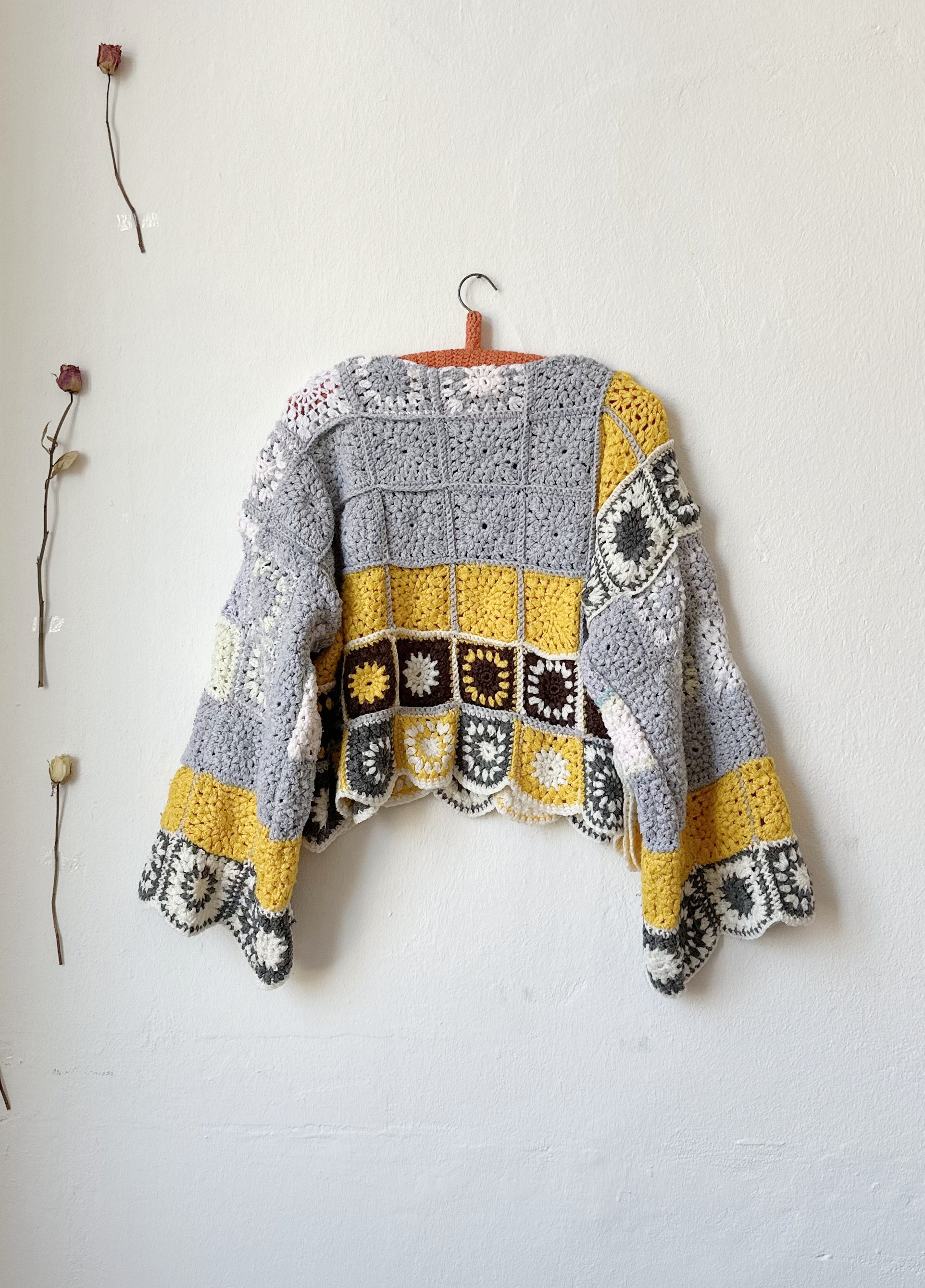 Back view of a crocheted sweater hanging on the wall. The sweater is grey and yellow crochet squares.
