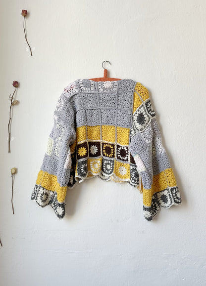 Back view of a crocheted sweater hanging on the wall. The sweater is grey and yellow crochet squares.