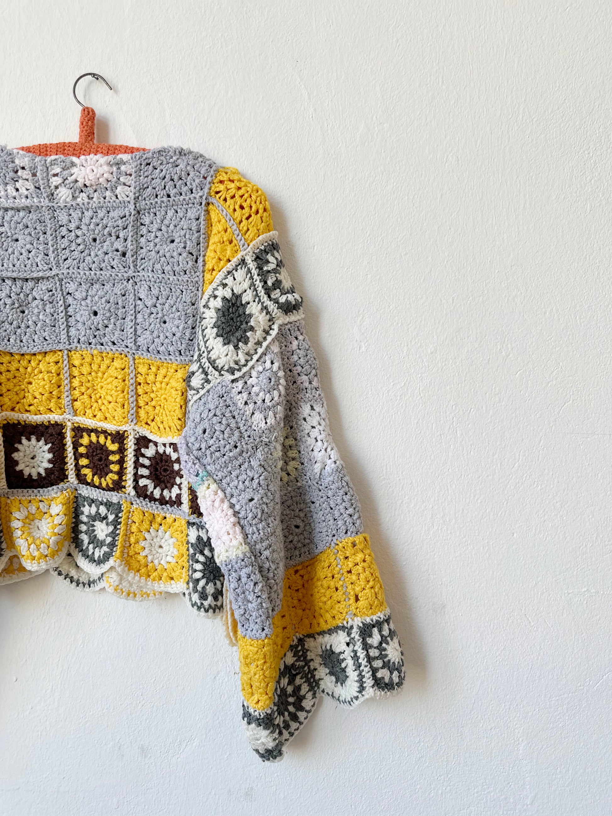 A detailed view of the back of a crocheted sweater hanging on the wall. The sweater is grey and yellow made from granny squares blanket.