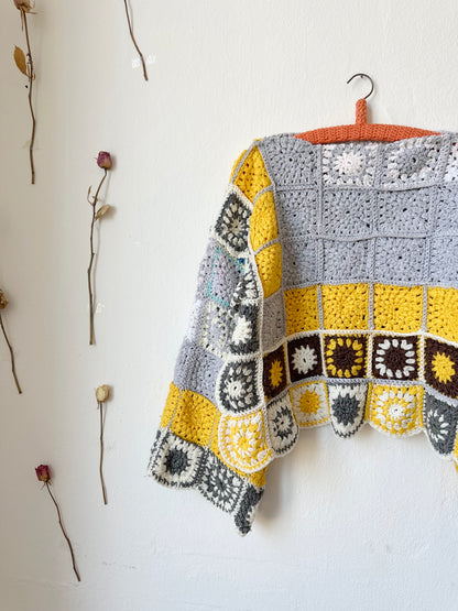 A detailed front view of a crocheted sweater hanging on a wall. The sweater is grey and yellow made from a granny square blanket.