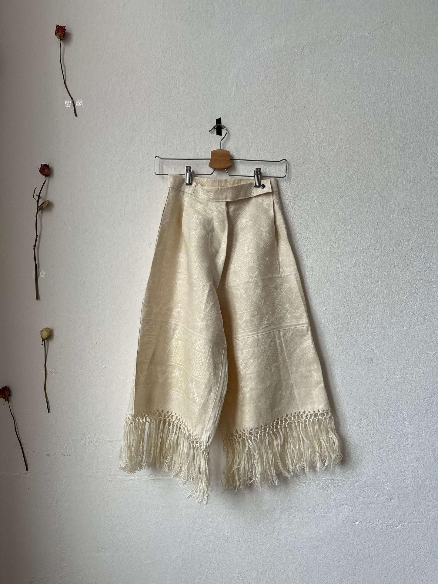 Wide leg, high-waisted trousers with tassels along the hem, displayed on a hanger against a plain wall.