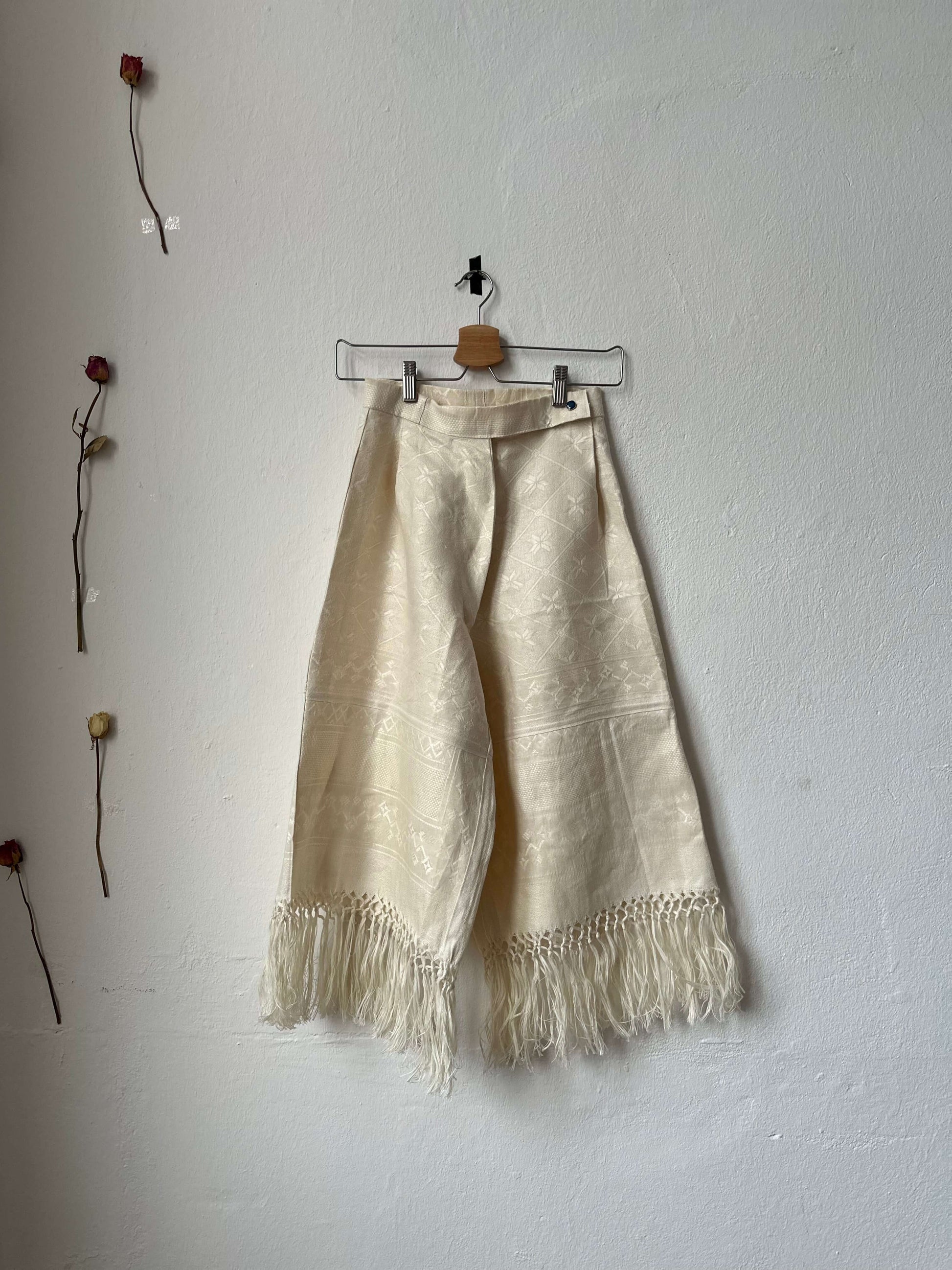 Wide leg, high-waisted trousers with tassels along the hem, displayed on a hanger against a plain wall.