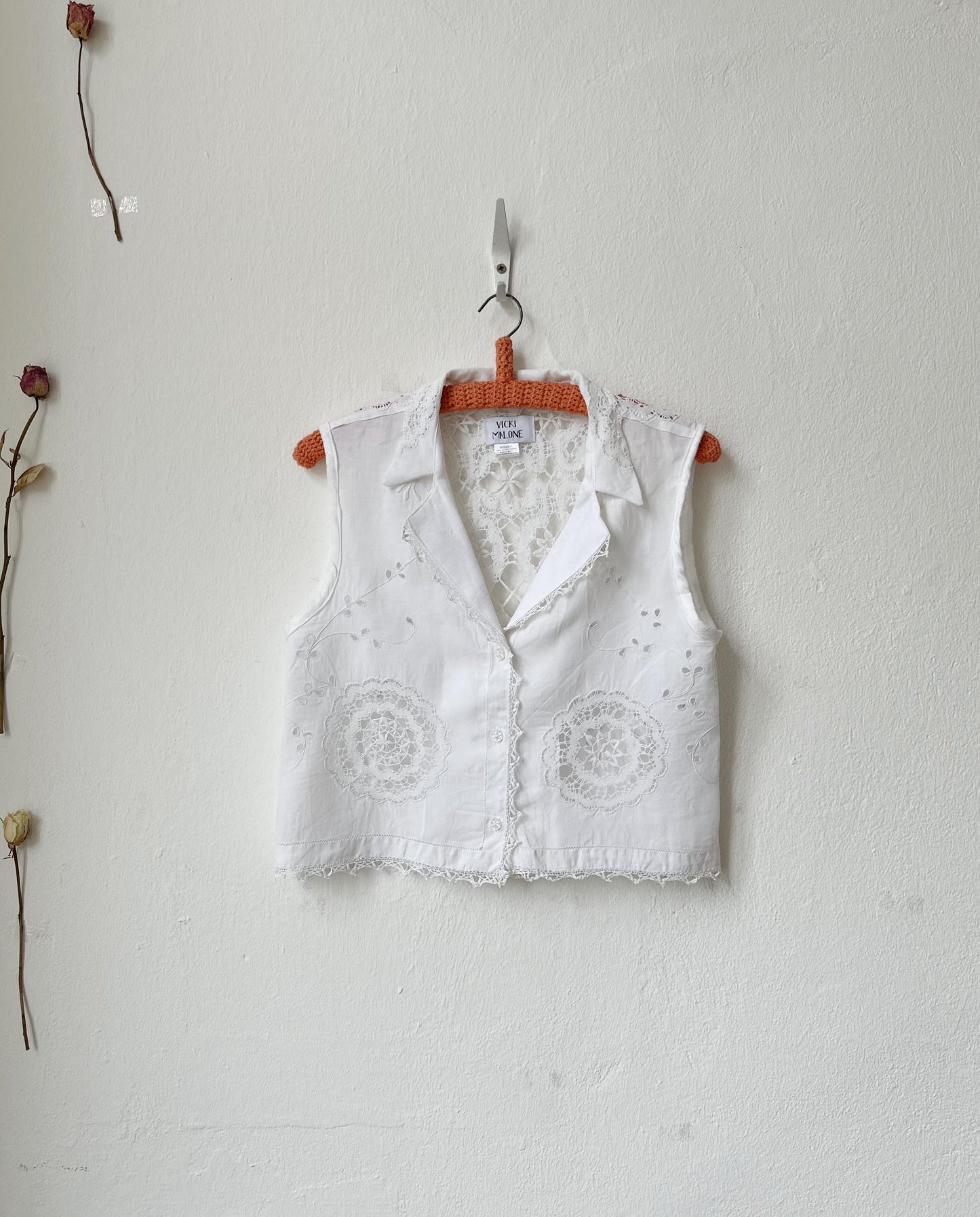 White embroidered vest hanging on a wooden hanger against a light gray wall.