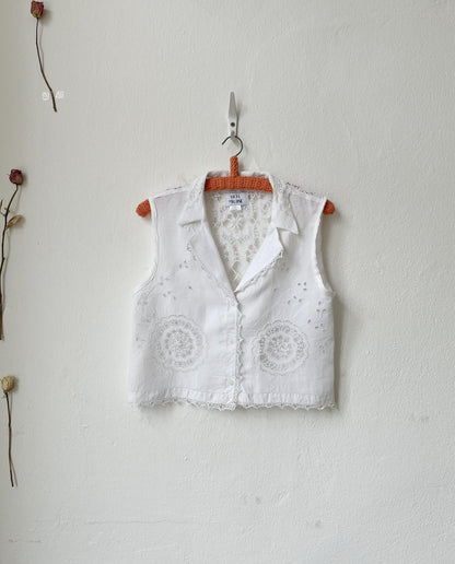 White embroidered vest hanging on a wooden hanger against a light gray wall.