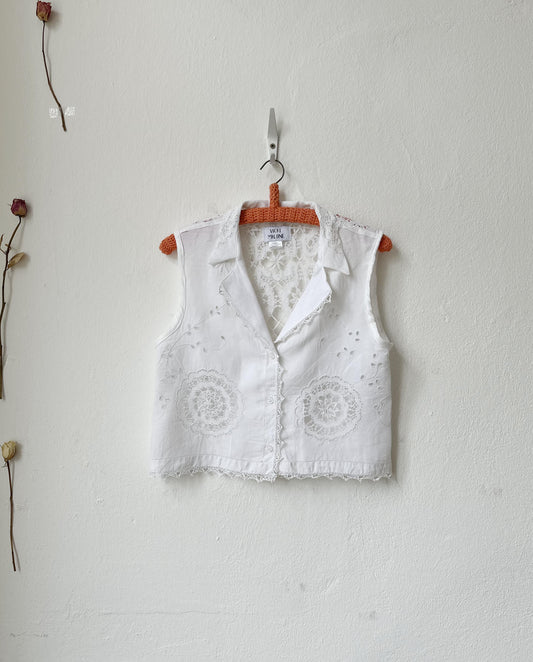 White embroidered vest hanging on a wooden hanger against a light gray wall.