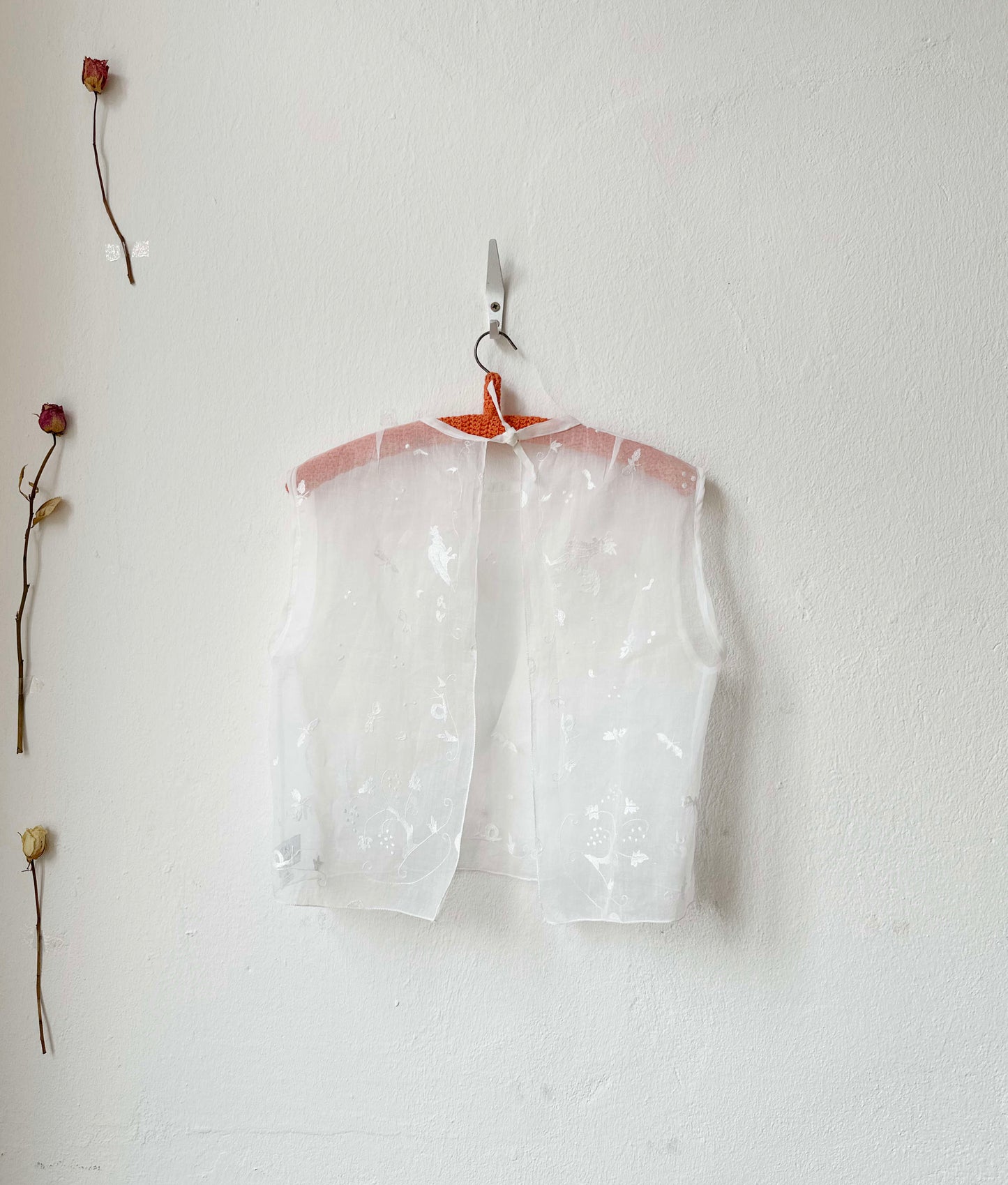Sheer embroidered vest top on a hanger against a white wall with dried flowers.