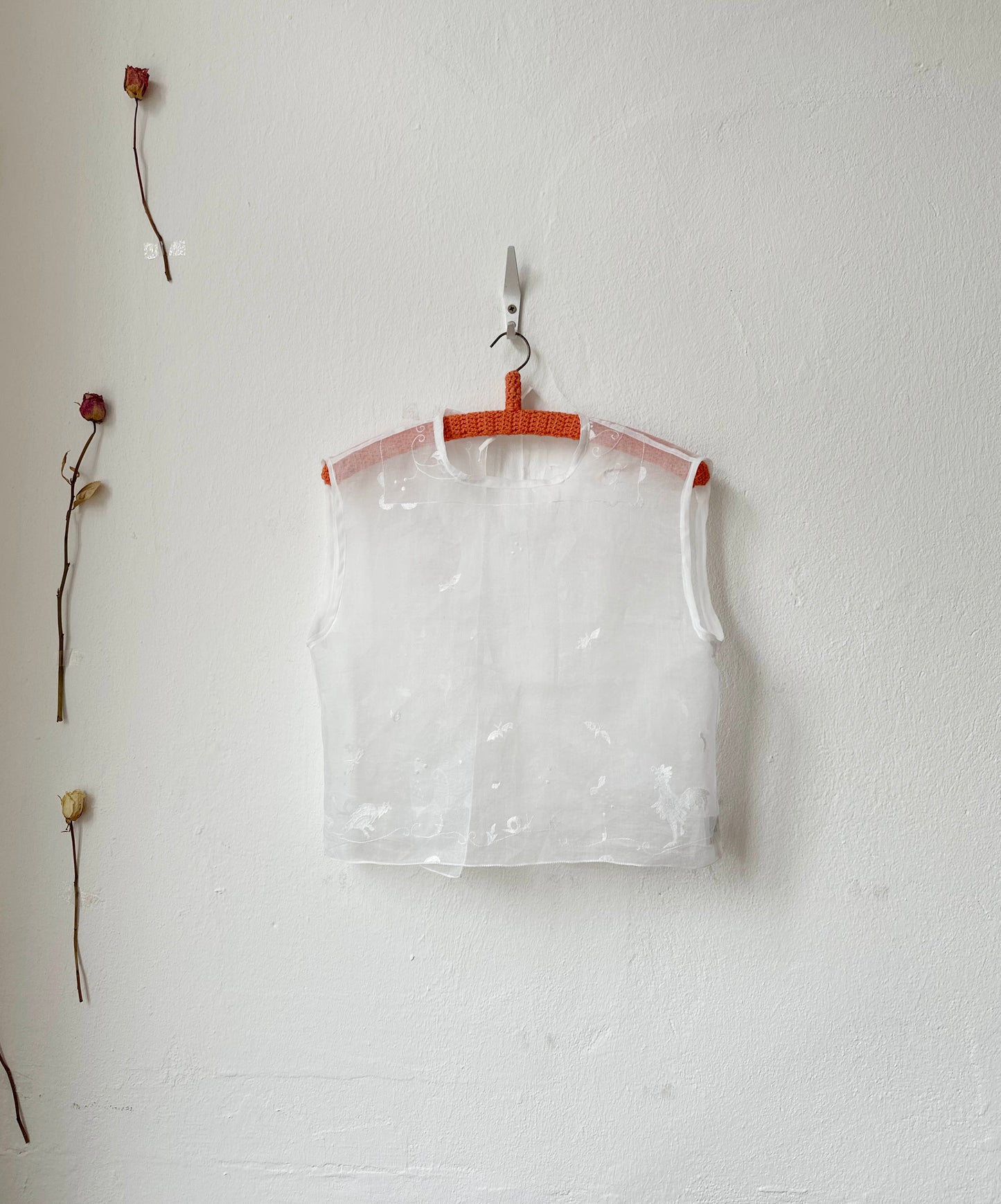 Sheer embroidered vest top hanging on a hook against a white wall with dried flowers.