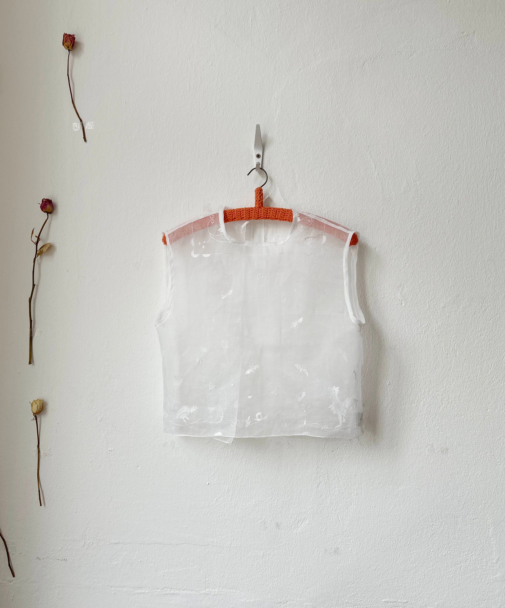 Sheer embroidered vest top hanging on a hook against a white wall with dried flowers.