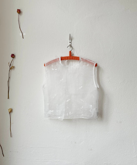 Sheer embroidered vest top hanging on a hook against a white wall with dried flowers.