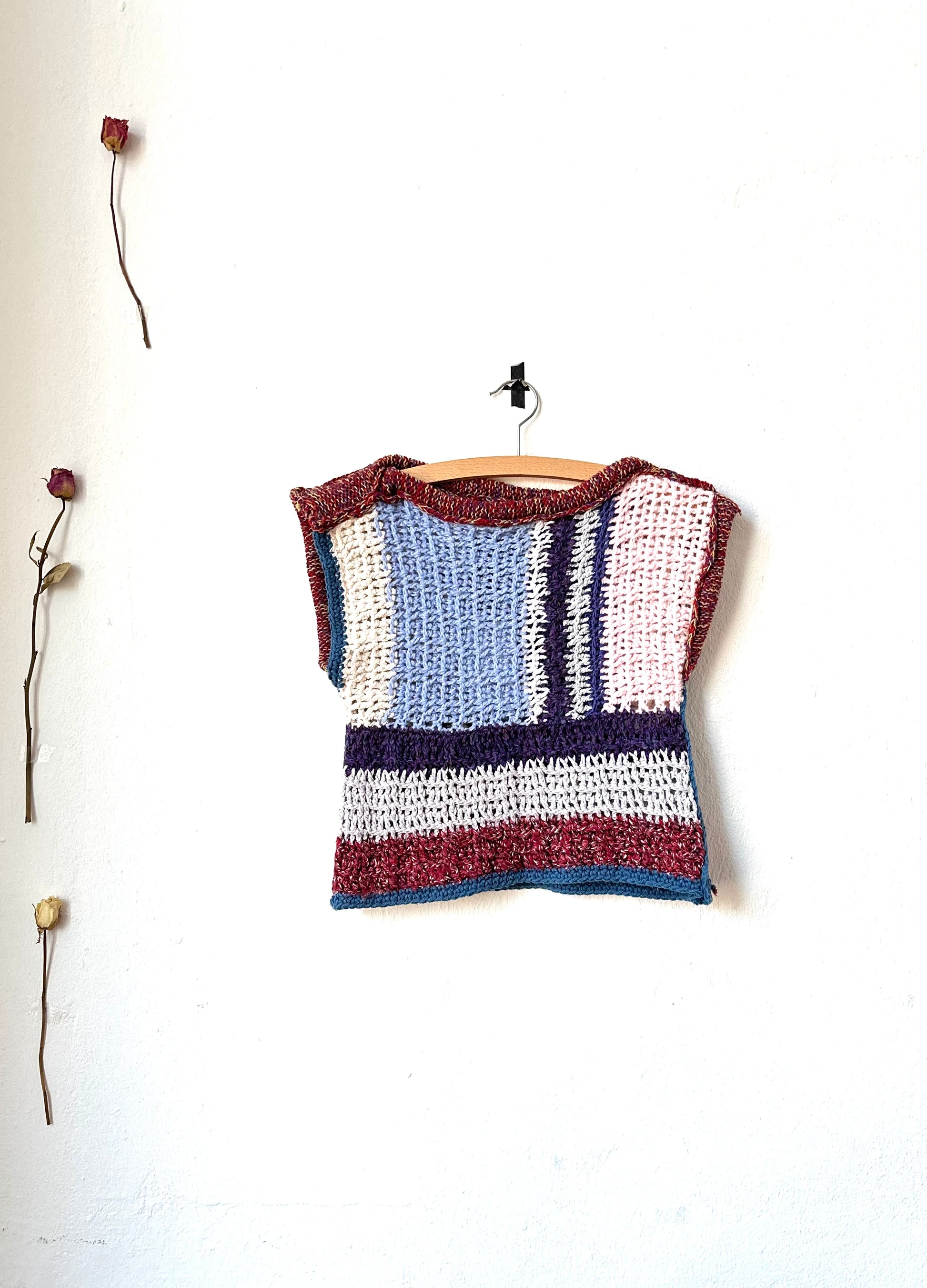 A hand crocheted blanket vest with a striped pattern, displayed on a wooden hanger against a white wall, with decorative branches and flowers below.
