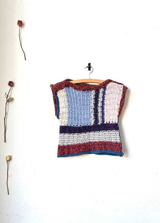 A hand crocheted blanket vest with a striped pattern, displayed on a wooden hanger against a white wall, with decorative branches and flowers below.