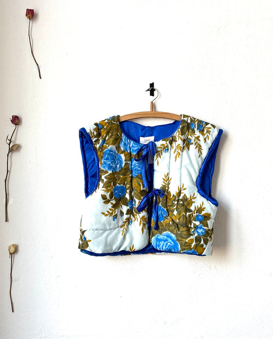 A blue quilted vest with a floral pattern displayed on a hanger against a white wall, with dried flowers attached to the wall.