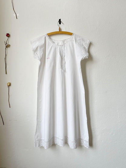 Vintage white dress with lace details hanging on a wooden hanger against a white wall.