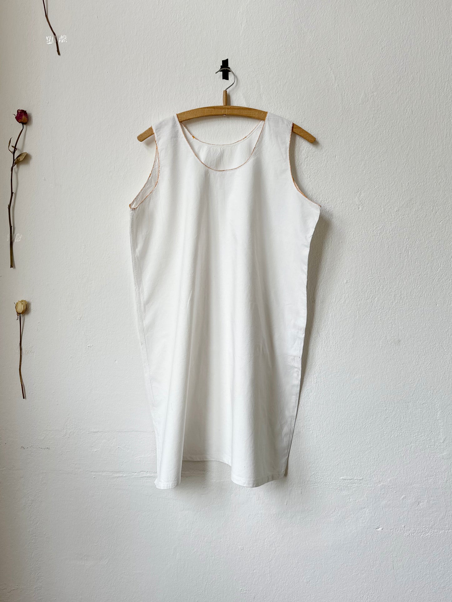 White sleeveless vintage dress with mustard stitching hanging on a wooden hanger against a plain white wall.