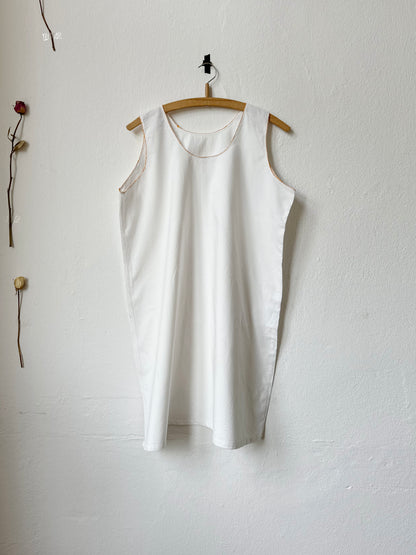 White sleeveless vintage dress with mustard stitching hanging on a wooden hanger against a plain white wall.