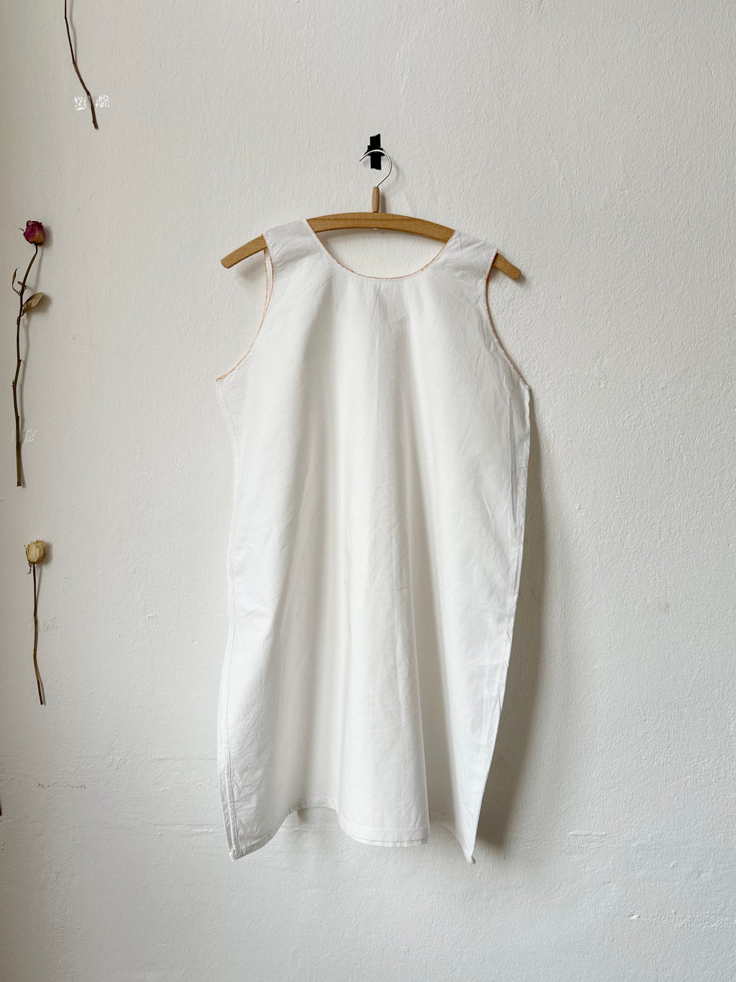Vintage white sleeveless dress hanging on a wooden hanger against a white wall with dried flowers.