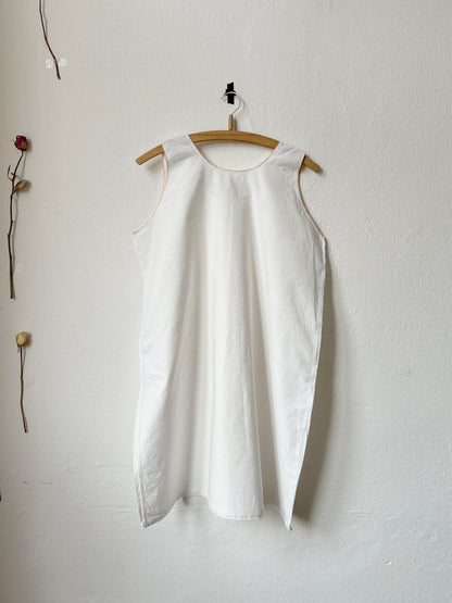 Vintage white sleeveless dress hanging on a wooden hanger against a white wall with dried flowers.