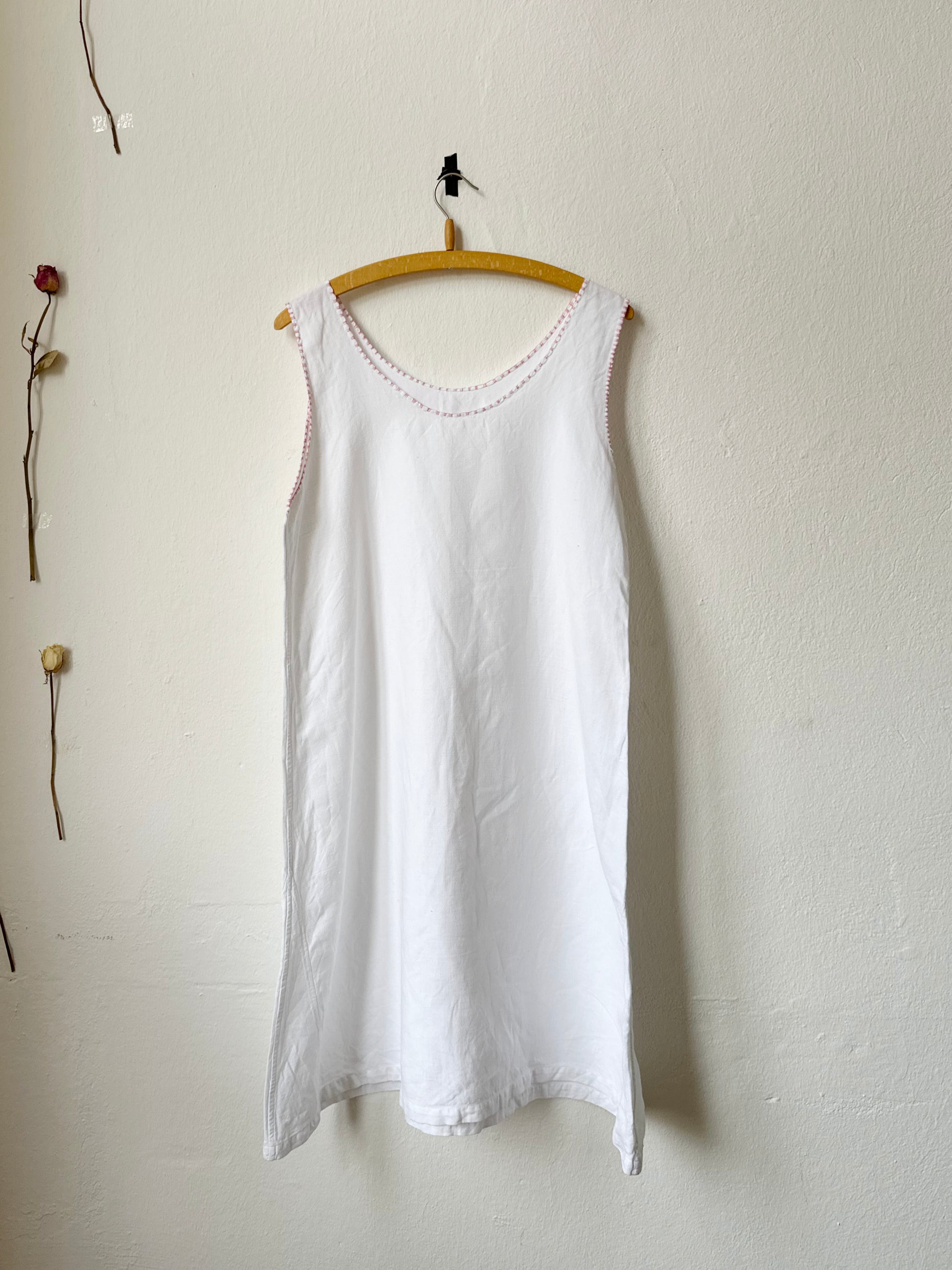 White sleeveless dress with pink trim hanging on a wooden hanger against a plain white wall.