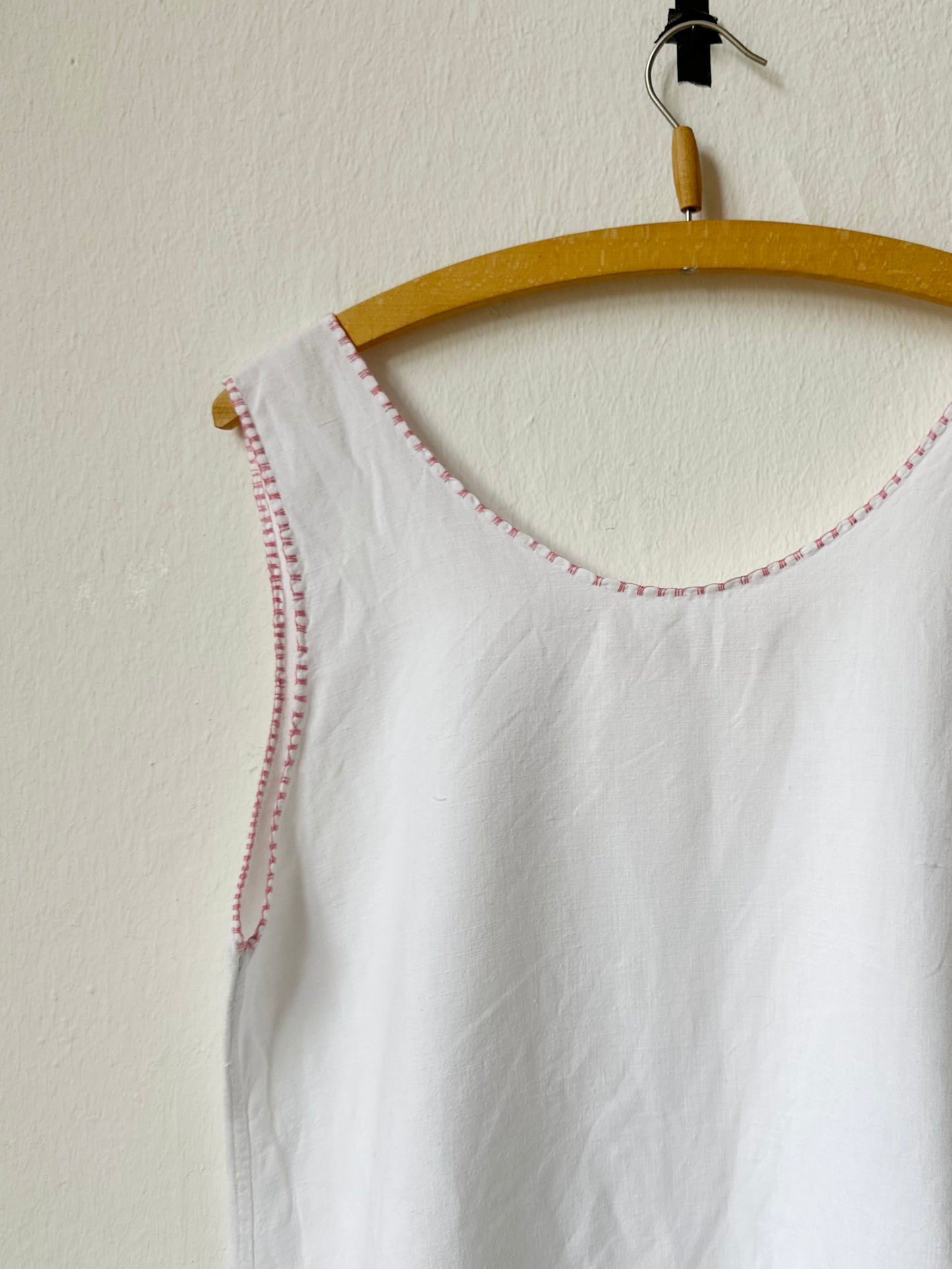 White sleeveless vintage dress with pink trim on a wooden hanger against a beige wall.