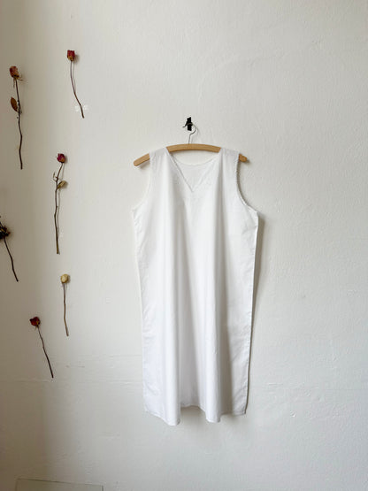 White sleeveless vintage dress hanging on a wooden hanger against a white wall with dried flowers.