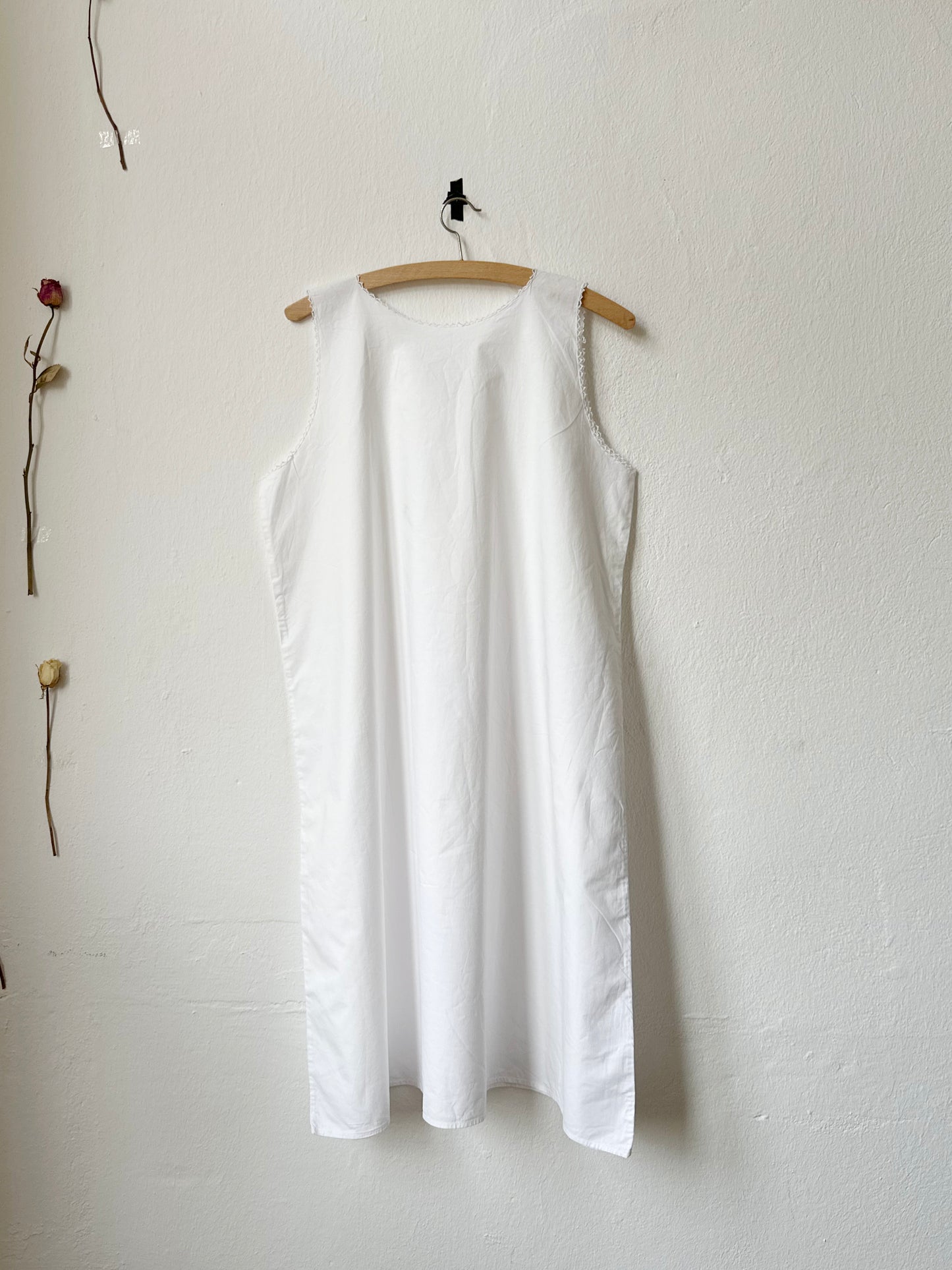 White sleeveless vintage dress hanging on a wooden hanger against a white wall.