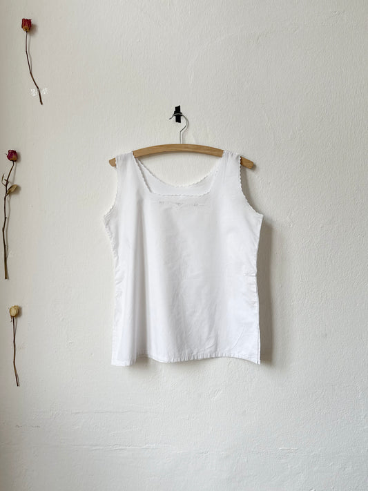 White tank top hanging on a wooden hanger against a plain white wall with two dried roses.