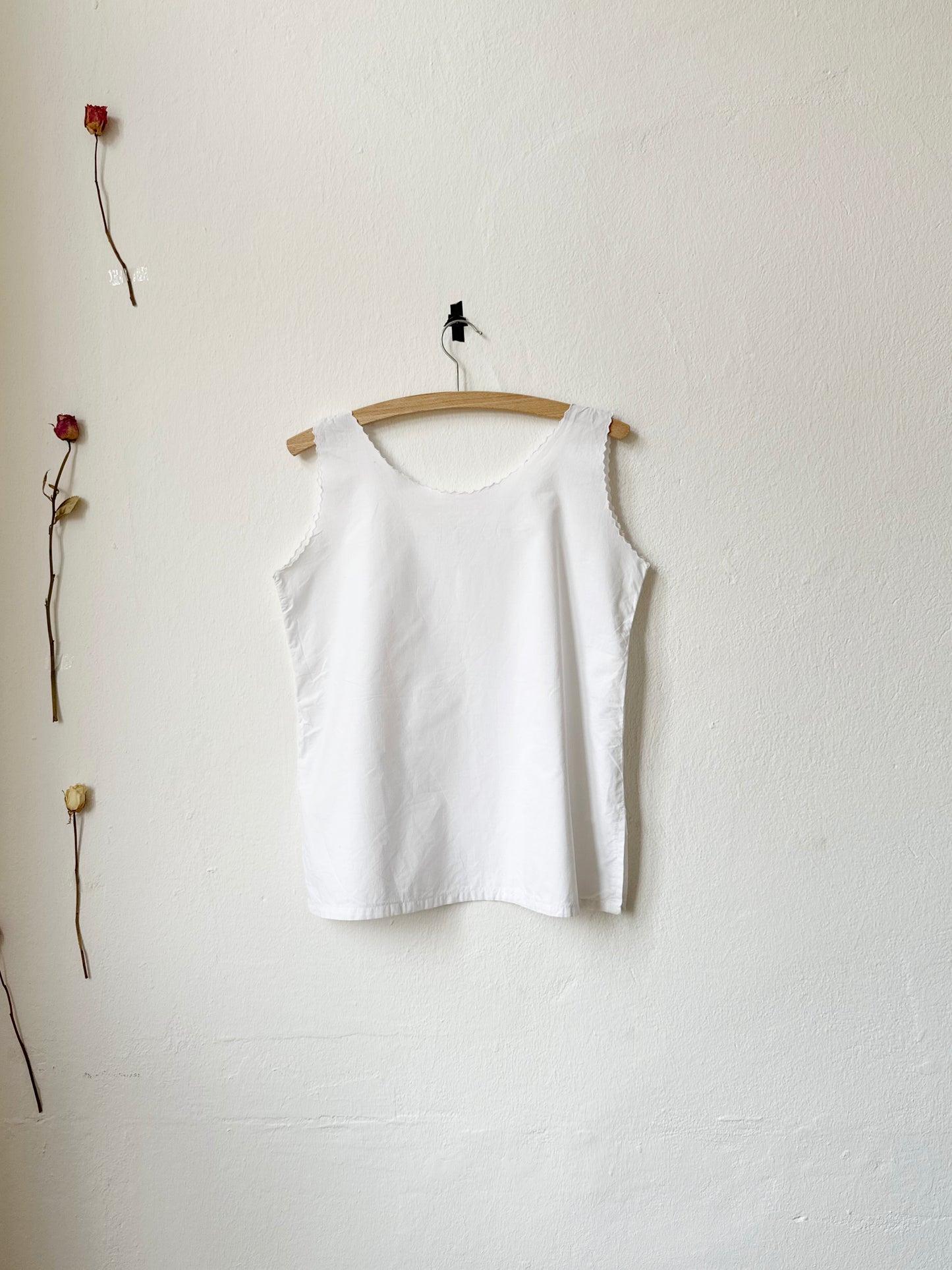 Vintage white tank top on a wooden hanger against a white wall with two dried roses.
