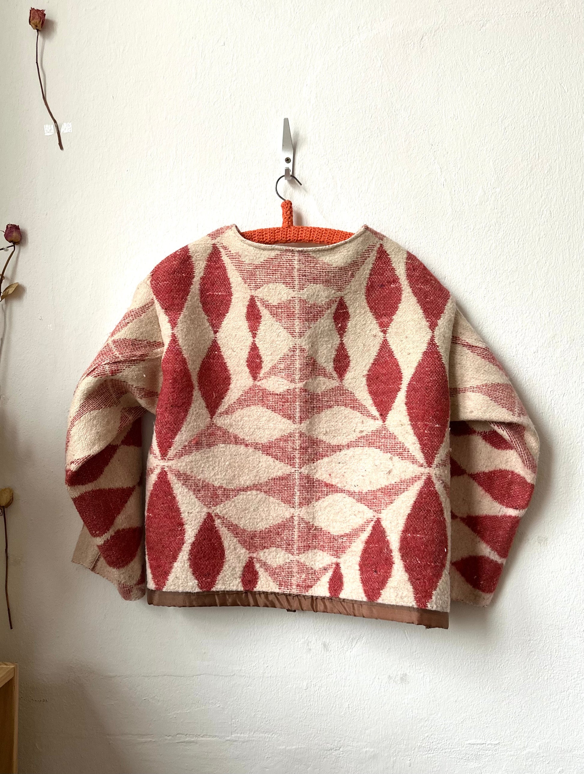 Red and beige patterned blanket jacket
 hanging on a hook against a white wall.