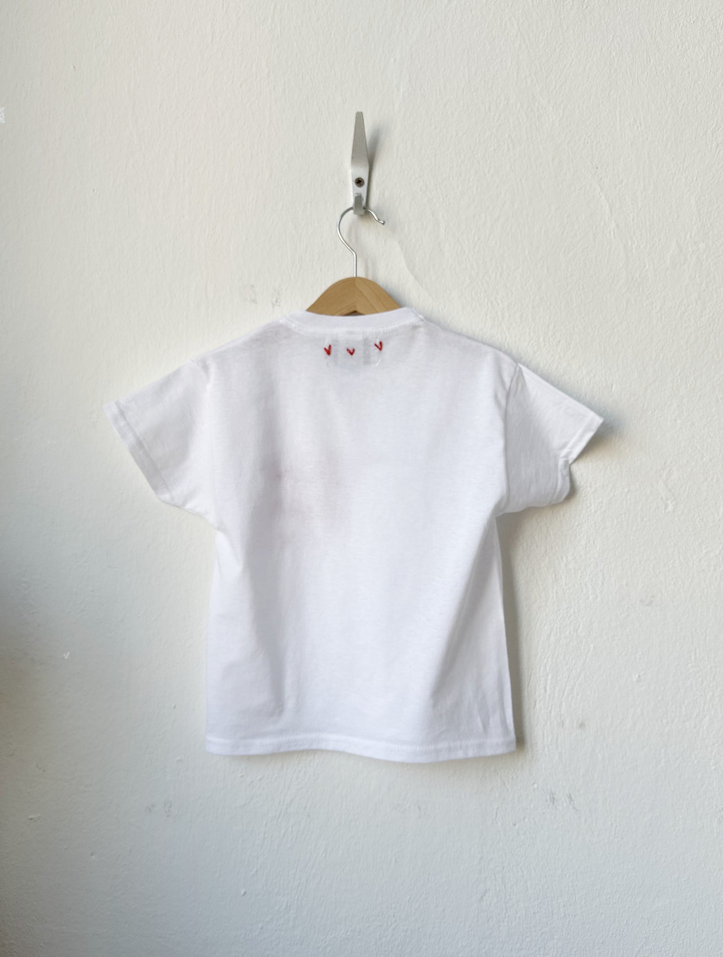 White t-shirt with small red hearts on a hanger against a white wall