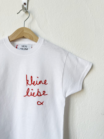 White t-shirt with red text 'kleine liebe' on a wooden hanger against a white wall.